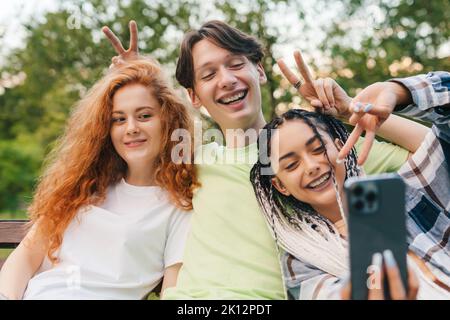 Vue de face de trois amis assis sur un banc en bois dans un parc prenant un selfie à l'aide d'un dispositif moderne. Selfie de jeunes souriants qui s'amusent Banque D'Images