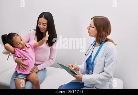 Belle fille coréenne assise à côté de sa mère au rendez-vous du pédiatre et montrant à son médecin son âge. Consultation, santé des enfants Banque D'Images