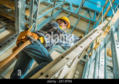 Travailleur de la construction avec un marteau suspendu à sa ceinture porte-outils en montant l'échelle d'extension pendant le travail sur la construction du cadre de squelette en acier du Hou Banque D'Images