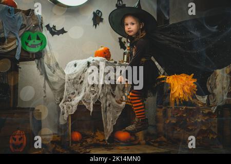 Petite fille de 3-4 ans en costume de sorcière et bas rayés vole sur le balai dans la décoration de cour d'Halloween avec des citrouilles orange vif d'halloween et Banque D'Images