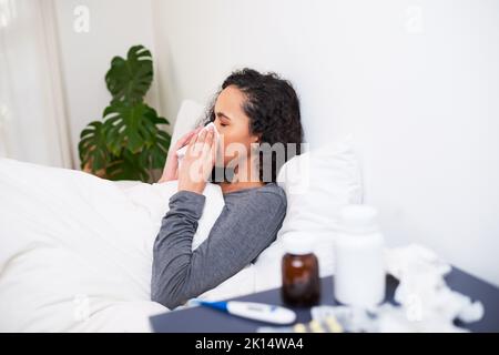 Une jeune femme multi-ethnique lui souffle le nez tout en se rétablissant de la grippe au lit Banque D'Images