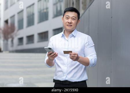 Portrait d'un jeune asiatique dans une chemise blanche, debout dans la rue près d'un bureau moderne, tenant un téléphone et une carte de crédit, regardant l'appareil photo. Banque D'Images