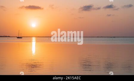 Un disque jaune de soleil se levant dans un ciel orange, au-dessus d'une mer Méditerranée calme, reflété dans l'eau à côté d'un seul voilier Banque D'Images