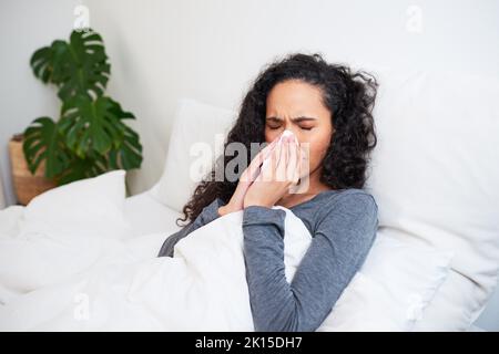 Une jeune femme multi-ethnique lui souffle le nez tout en se rétablissant de la grippe au lit Banque D'Images