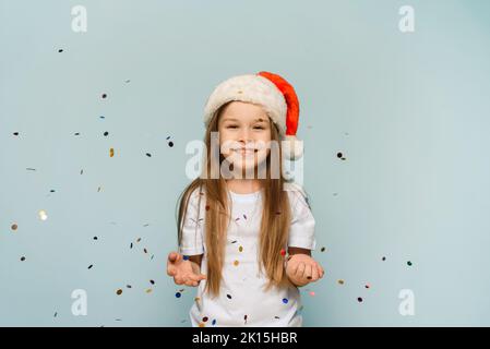 Petite fille mignonne dans le chapeau de père noël jette des confetti et regarde l'appareil photo. fête de noël. Banque D'Images