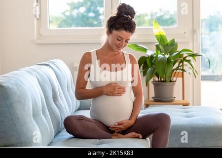 Jolie femme enceinte assise sur un canapé et tenant le ventre avec tendresse et amour Banque D'Images