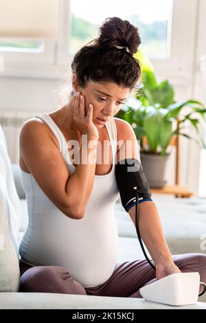 Femme enceinte inquiète vérifiant la tension artérielle et le pouls à la maison avec le dispositif de fréquence cardiaque Banque D'Images