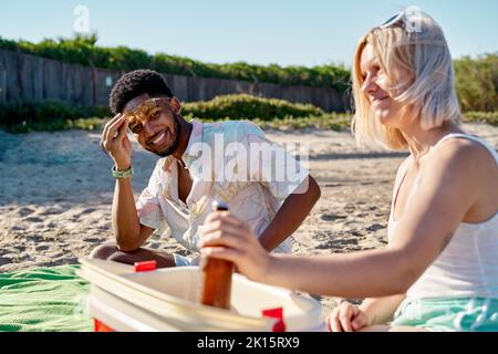 Joyeux Afro-américain regardant l'appareil photo avec le sourire et en ajustant les lunettes de soleil sur la tête tout en étant assis près de la femme prenant de la bière de glacière pendant le picni Banque D'Images