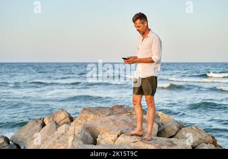 Homme d'affaires plein corps pieds nus mature en chemise et short blanc debout sur des pierres près de la mer et de la navigation sur les médias sociaux sur smartphone l'été W. Banque D'Images