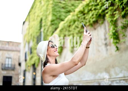 Vue latérale d'une touriste féminine en lunettes de soleil et un chapeau de soleil se prenant sur un smartphone près du bâtiment en pierre avec de l'ivy pendant les vacances d'été Banque D'Images