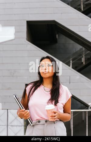 Jeune femme entrepreneure de l'ethnie pensive avec de longs cheveux foncés dans des vêtements décontractés et des lunettes qui regardent loin en se tenant sur la rue de la ville avec la coupe o Banque D'Images