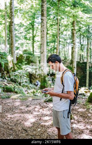 Homme dans des vêtements décontractés avec sac à dos parcourant la carte GPS sur un téléphone portable tout en explorant la forêt verte le jour ensoleillé de l'été Banque D'Images