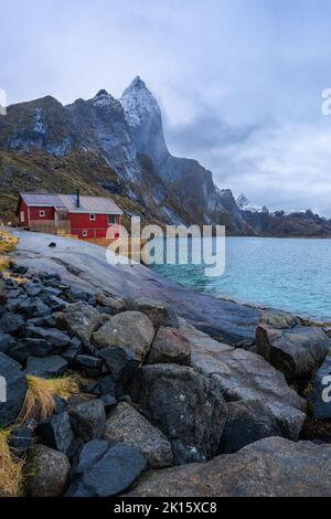 Paysage marin pittoresque avec maison rouge sur la côte rocheuse et montagnes massives avec des sommets enneigés sous ciel nuageux en Norvège Banque D'Images