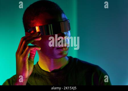 Jeune Afro-américain concentré en t-shirt noir ajustant les lunettes futuristes modernes tout en faisant l'expérience de la réalité virtuelle en studio avec la ne colorée Banque D'Images