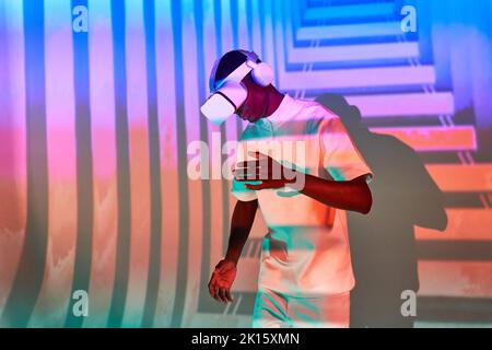 Jeune homme afro-américain élégant millénaire en t-shirt blanc expérience de la réalité virtuelle dans un casque VR moderne en studio dansant au néon coloré Banque D'Images