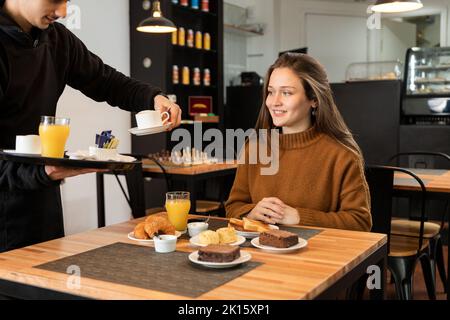 Jeune homme écourté, non reconnaissable, en uniforme noir, prenant un verre de jus du plateau tout en servant des plats pour femme souriante pendant le petit-déjeuner au restaurant Banque D'Images