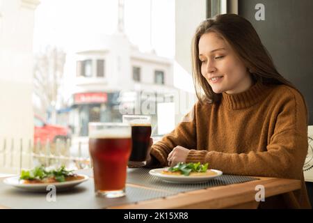 Jeune femme joyeuse en chandail brun souriant et dégustant une bière foncée tout en étant assise à la table du restaurant près de la fenêtre Banque D'Images