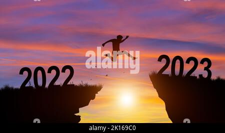 Joyeux Noël et bonne année en 2023. Homme sautant à travers l'écart de 2022 à 2023 falaise avec coucher de soleil et ciel crépuscule arrière-plan. Banque D'Images