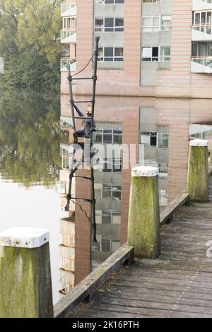 Apeldorn - Une scène paisible au bord de la rivière avec une promenade en bois, un bâtiment réfléchissant dans l'eau et une sculpture en métal. Banque D'Images