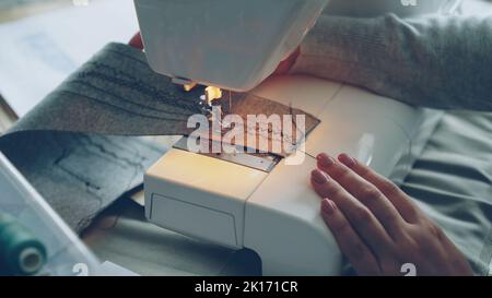 Vue rapprochée de la machine à coudre électrique moderne qui travaille la couture d'un morceau de tissu. La main manucurée de la jeune fille et les fils de couture colorés sont visibles. Banque D'Images