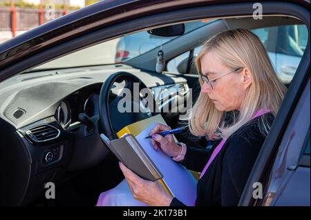 femme d'affaires d'âge moyen dans des lunettes en voiture travaillant avec des documents, concept de technologie mobile Banque D'Images