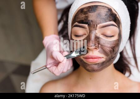 cosmetologiste appliquant le masque noir sur le visage jolie femme portant des gants noirs, femme magnifique au spa ayant des procédures faciales Banque D'Images