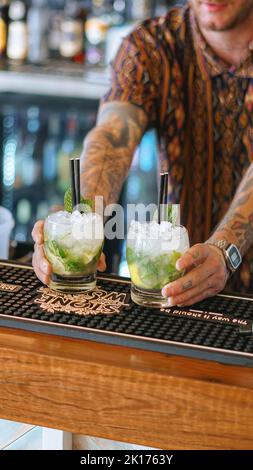 Un barman de sexe masculin sert 2 cocktails Mojito depuis l'arrière du bar. Il porte une chemise noire, une montre et sert les boissons avec deux pailles noires. Banque D'Images