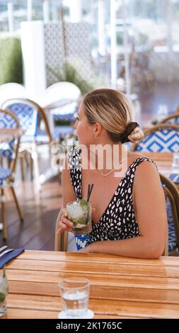 Une fille blonde appréciant un cocktail mojito, elle porte une robe à pois noir et blanc. Elle regarde loin car elle tient le cocktail. Banque D'Images