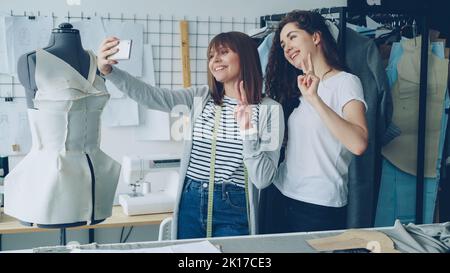 Deux femmes gaies stylistes de vêtements font drôles selfie avec smartphone tout en se tenant à côté de mannequin vêtu en studio. Les femmes attrayantes sont souriantes et posent. Banque D'Images