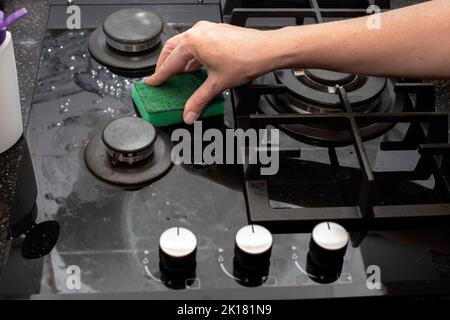 Une femme au foyer nettoie la cuisinière noire en vitrocéramique à gaz de la cuisine avec une éponge. Nettoyage de la maison. Banque D'Images