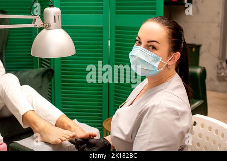 Jeune podiatre femelle avec un masque protecteur faisant de la chiropodie et regardant la caméra dans sa clinique de podiatrie Banque D'Images