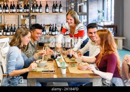 Des amis variés et positifs mangeant de délicieux plats à table avec des verres de vin rouge pendant le dîner dans un restaurant moderne et lumineux Banque D'Images