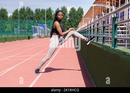 African American sportswoman étire les jambes sur la piste de jogging se préparant à courir le jour ensoleillé Banque D'Images