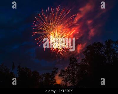 Feux d'artifice orange qui explosent au-dessus des arbres avec de la fumée et des nuages dans le ciel. Banque D'Images