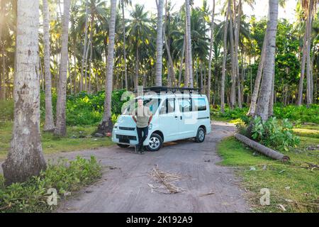Un jeune homme heureux debout devant une camionnette de camping-car blanche garée sur une route de terre dans un champ tropical de cococotiers à Bali Indonésie au coucher du soleil Banque D'Images