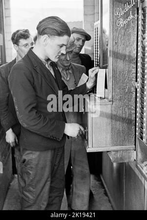 Années 1930, historiques, travailleurs de scierie arrivant au travail à British Coated Board & Paper Mills Ltd, Galles du Sud, Royaume-Uni, utilisant un enregistreur de temps Gledhill Brook avec un boîtier en chêne. La photo montre les travailleurs qui font la queue, avec un jeune homme qui utilise une clé et une carte sur la machine à cadencer. Une société britannique, basée à Huddersfield, Gledhill Brooks a produit une gamme d'enregistreurs de temps de haute qualité jusqu'à ce que l'entreprise soit reprise en 1964. Banque D'Images