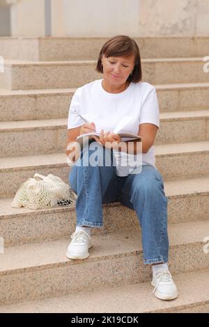 femme mature travaille à distance, l'enseignement supérieur pour les adultes, femme écrit et note dans un carnet assis sur les escaliers à l'extérieur, indépendant. Collège s Banque D'Images