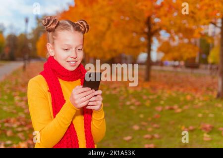 une écolière dans un chandail orange et un foulard rouge tient un téléphone contre le fond des arbres jaunes d'automne et sourit. Banque D'Images