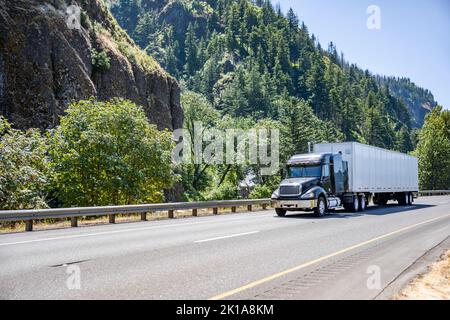 Tracteur semi-remorque industriel noir frais transportant des marchandises commerciales dans une remorque semi-remorque à camion sec conduite sur la route avec la montagne et la forêt i. Banque D'Images