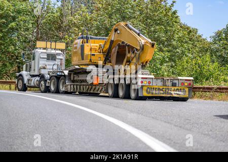 Tracteur semi-remorque blanc à cabine de jour industriel avec signalisation surdimensionnée transportant la pelle hydraulique en montée dans les virages, conduite sur route étroite sur t Banque D'Images