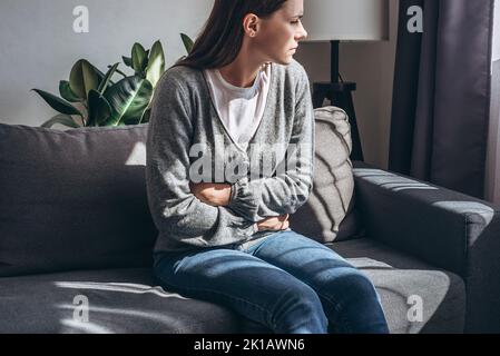 Jeune femme malheureuse 20s ans assis sur un canapé dans la salle de séjour à la maison, souffrant de douleurs abdominales crampes, respiration intense, holdi encombrant Banque D'Images