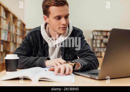Un étudiant est assis sur un bureau et regarde un ordinateur portable. Jeune homme en bibliothèque préparant des examens. Banque D'Images