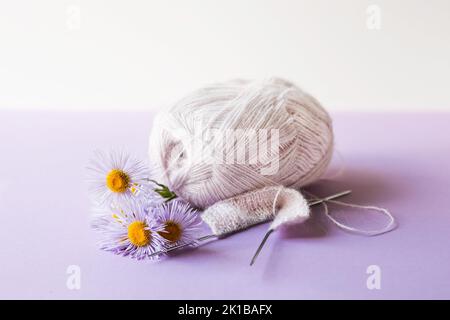 Une boule de fil rose tendre avec des aiguilles à tricoter et des fleurs d'erigeron pourpres sur fond violet Banque D'Images