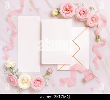Cartes en papier et enveloppe entre les roses rose clair et rubans de soie sur la vue de dessus rose, maquette de mariage. Scène romantique avec carte verticale et flo pastel Banque D'Images