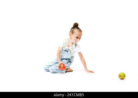 Jolie fille vêtue de combinaisons en denim qui se berce avec des pommes sur fond blanc. Banque D'Images