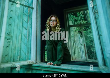 Une belle jeune femme blonde dans un chandail vert vintage se tient à la vue d'une fenêtre cassée dans une vieille maison abandonnée. Solitude. Banque D'Images