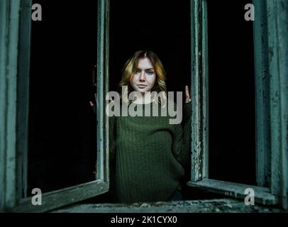 Une belle mystérieuse jeune femme blonde dans un chandail vert vintage se tient à une fenêtre cassée dans l'obscurité d'une ancienne maison de village abandonnée. Mon Banque D'Images