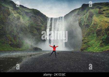 Jeune touriste en blouson rouge appréciant la cascade de Skogafoss coulant de la falaise en été en Islande. Place islandaise célèbre, destination de voyage Banque D'Images