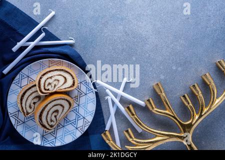 Assiette de désert Hanukkah avec rouleau de noix, bougies blanches, et menorah dorée sur fond bleu texturé Banque D'Images