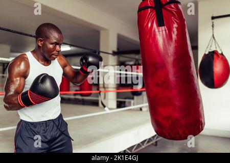 Boxer jeune et concentré s'entraîner avec un sac de poinçonnage rouge à la salle de gym. Jeune homme sportif pratiquant ses techniques de poinçonnage dans un gymnase de boxe. Banque D'Images